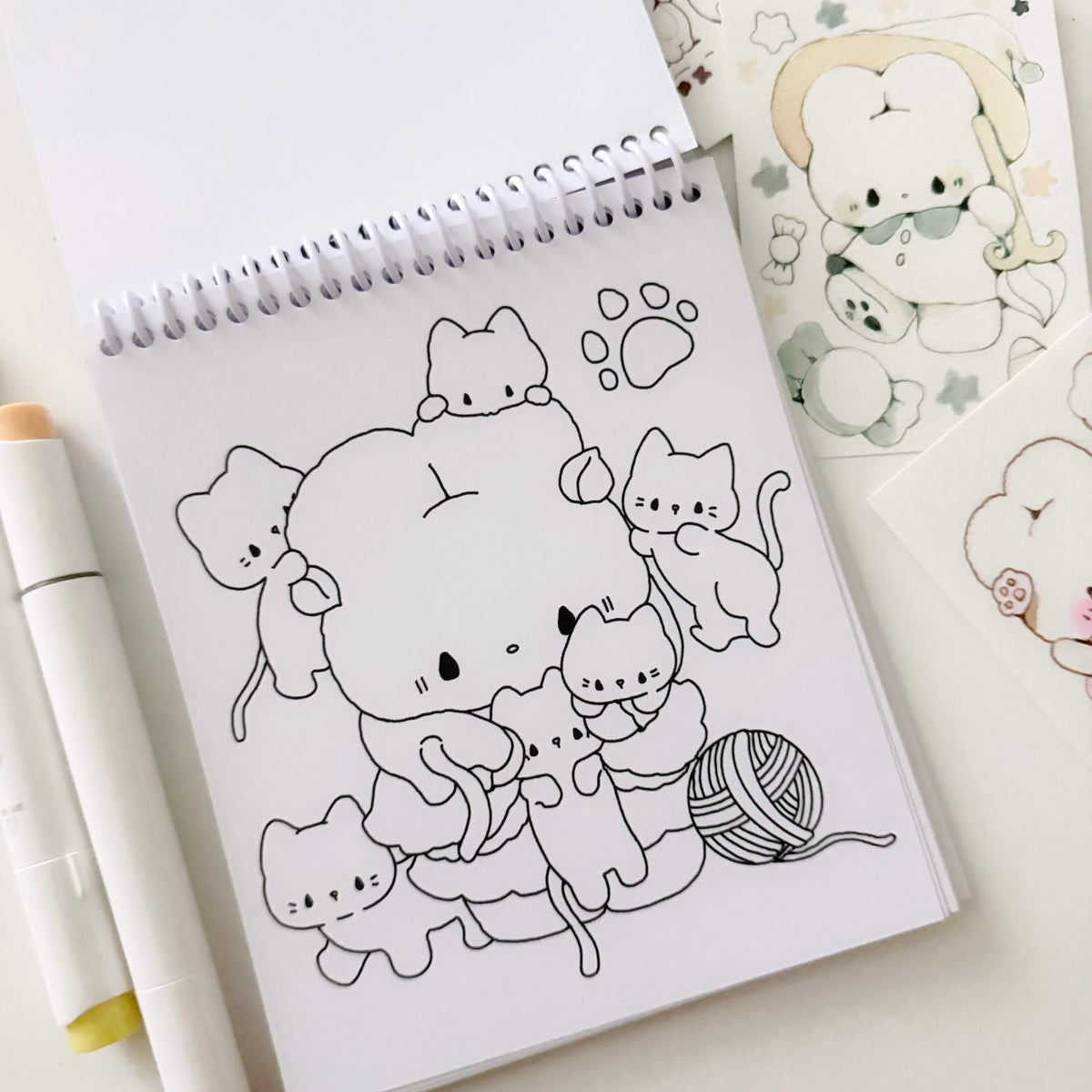 A spiral-bound coloring book or sketchpad with black line art of a cartoon character and several cats, shown on a light surface with a marker and other illustrated sheets nearby.