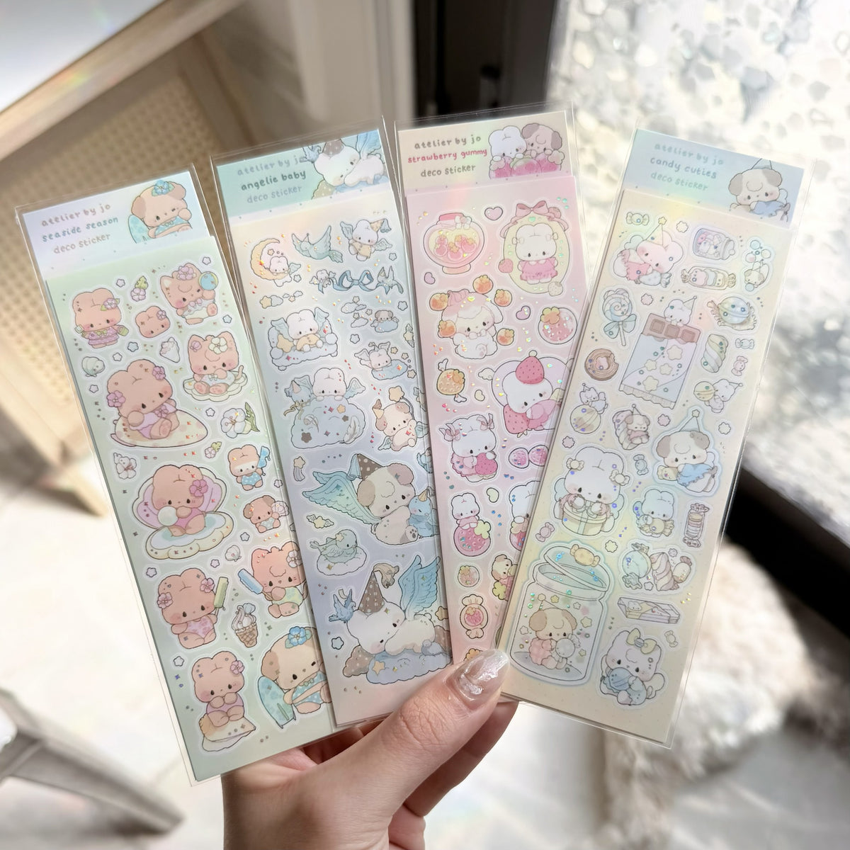 Four sheets of cute cartoon stickers held in a hand, featuring pastel-colored characters and objects on a softly blurred indoor background.