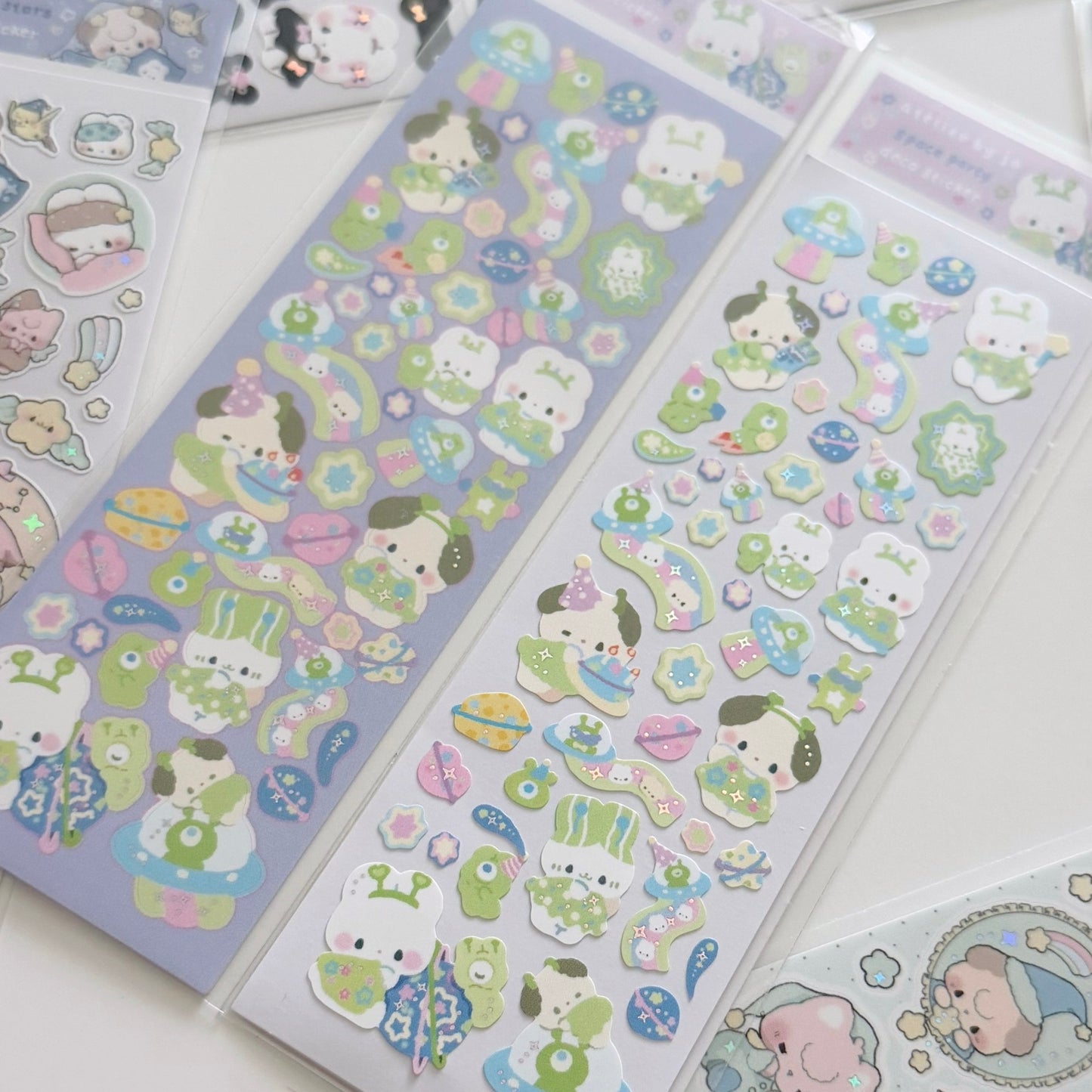 Three packaged sheets of cute cartoon stickers laid out on a white surface. The stickers feature pastel-colored characters and motifs including bunnies, frogs, flowers, stars, and small illustrated objects in soft purple, green, pink, blue, and white tones.