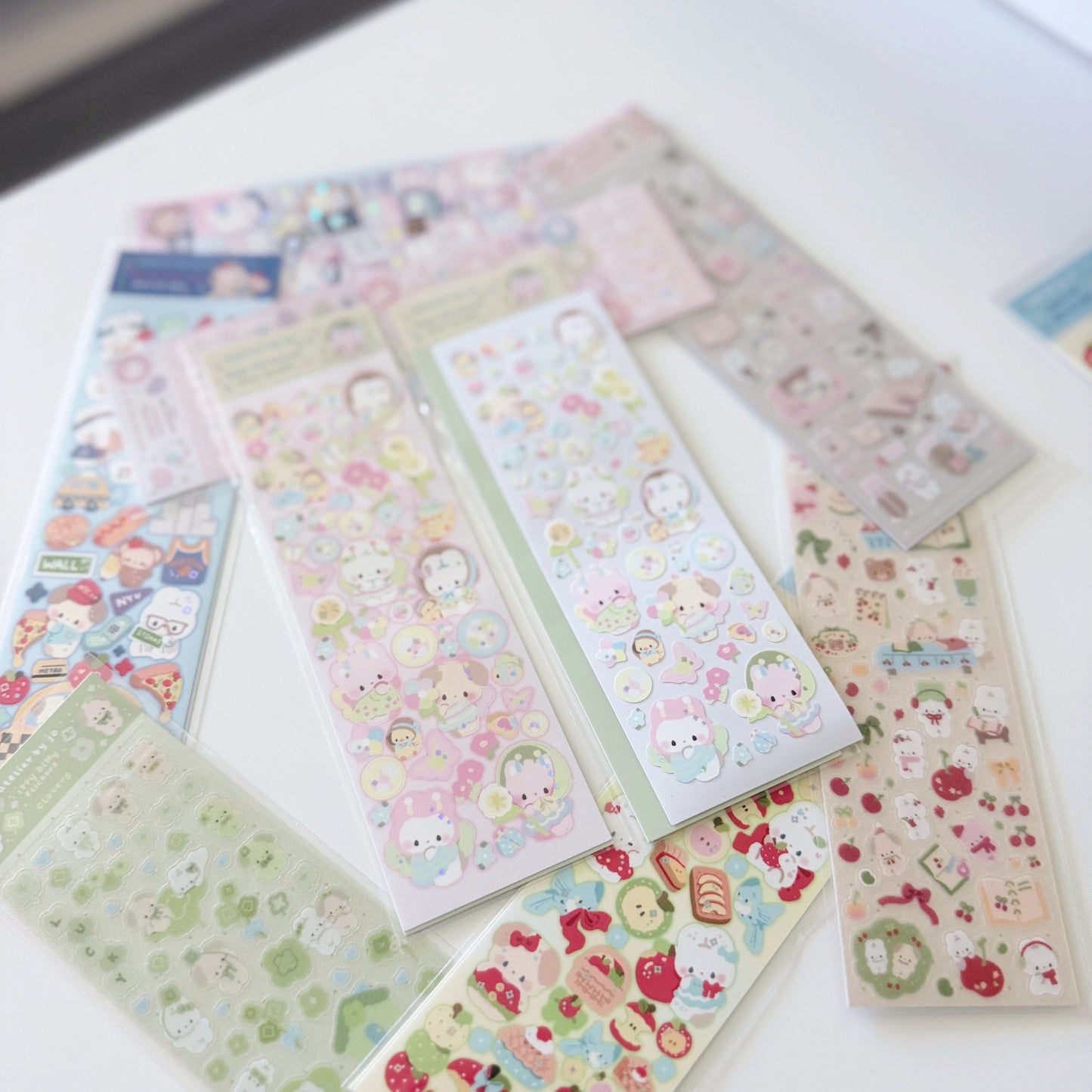 Several sheets of cute cartoon stickers arranged on a white surface. The stickers feature pastel-colored characters and illustrations, including strawberries, desserts, and small animals, in clear plastic sleeves.