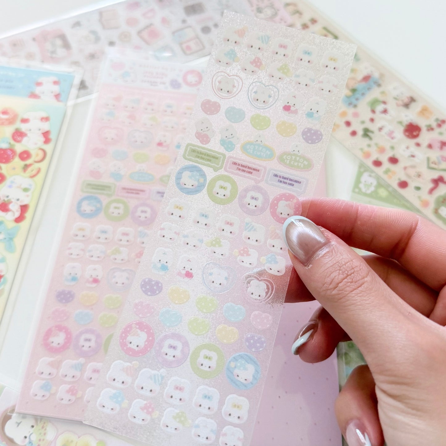 A hand with silver manicured nails holds a sheet of cute pastel stickers, with other sticker sheets visible in the background.