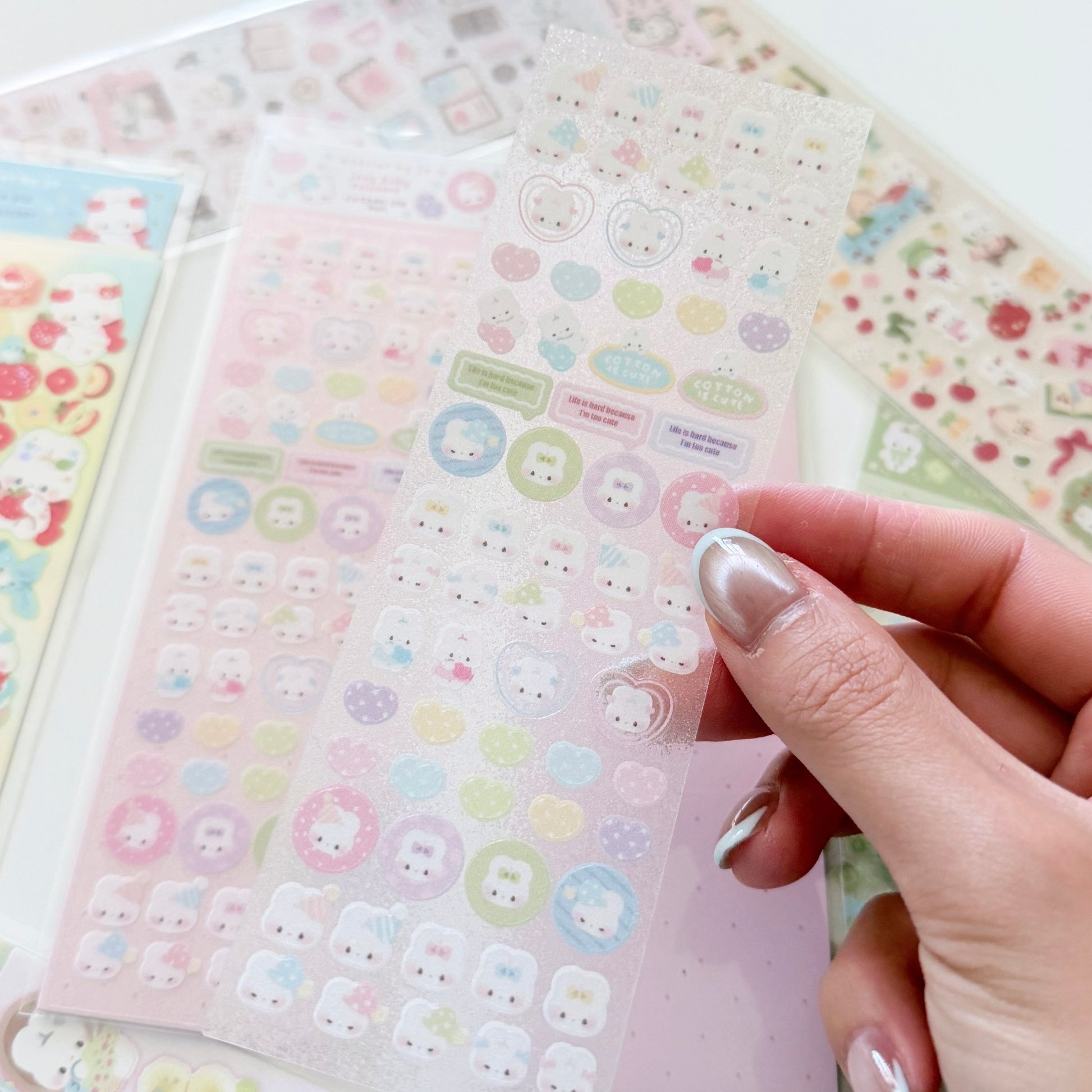 A hand with silver manicured nails holds a sheet of cute pastel stickers, with other sticker sheets visible in the background.