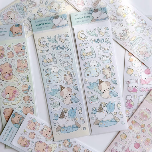 Image shows a white surface covered with sticker sheets in various pastel colors. The main focus are two blue sticker sheets with holographic foil detailing. 