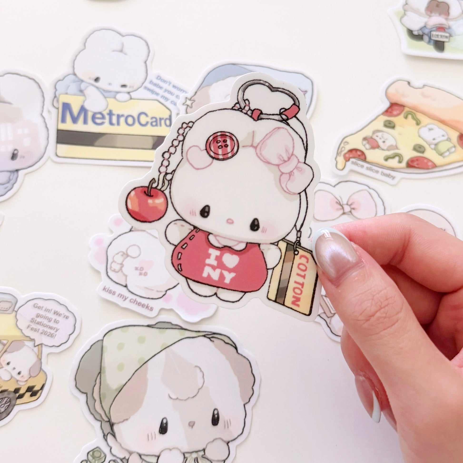 A hand is holding a die-cut sticker of a bunny with a red heart dress and 'I <3 NY' text, surrounded by other cute cartoon stickers on a white surface.