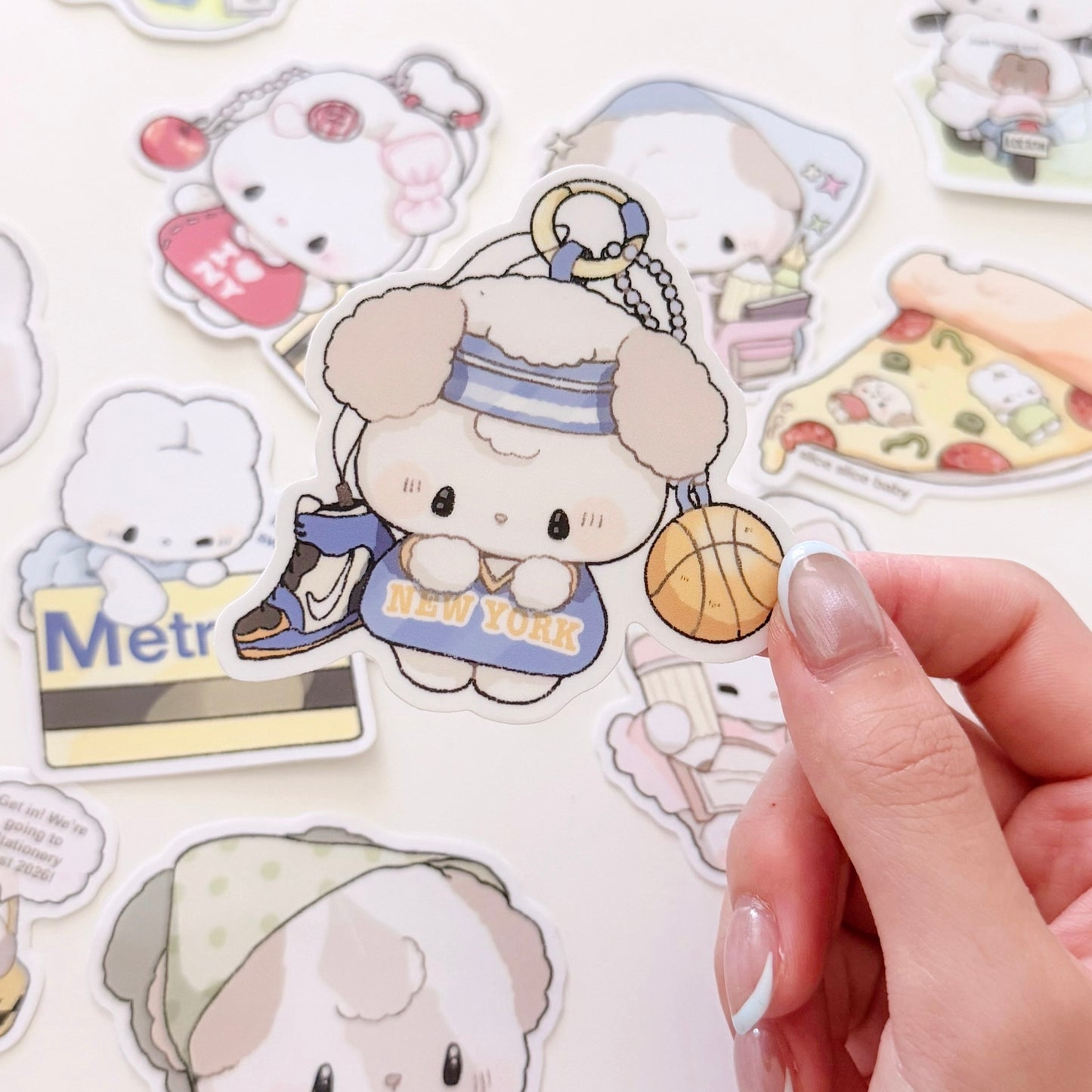 A close-up of a hand holding a die-cut sticker featuring a cute cartoon dog character in a blue New York-themed outfit with a basketball, surrounded by other colorful stickers on a white background.