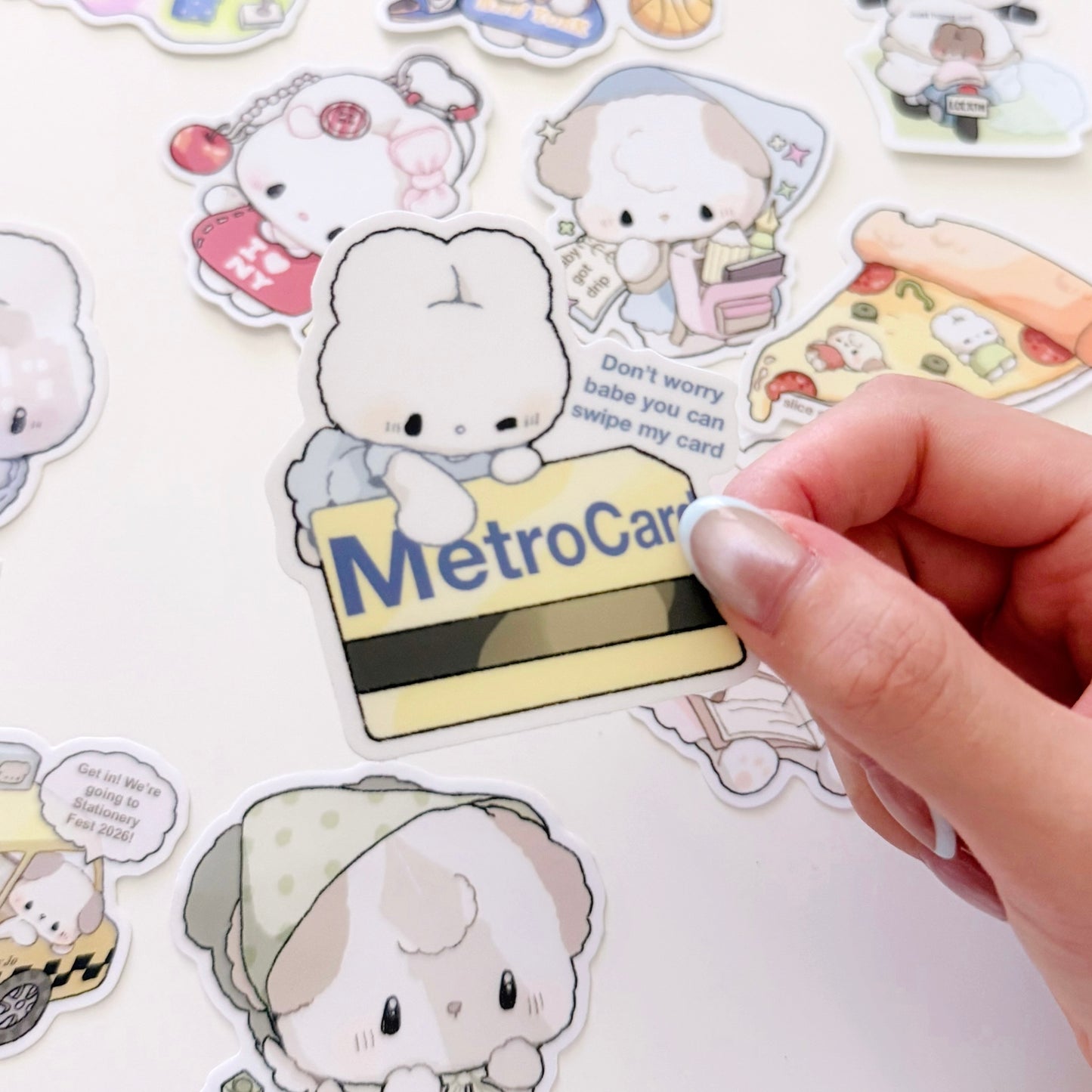 A hand is holding a die-cut sticker featuring a cute white bunny character with a MetroCard, surrounded by other pastel cartoon stickers on a white surface.