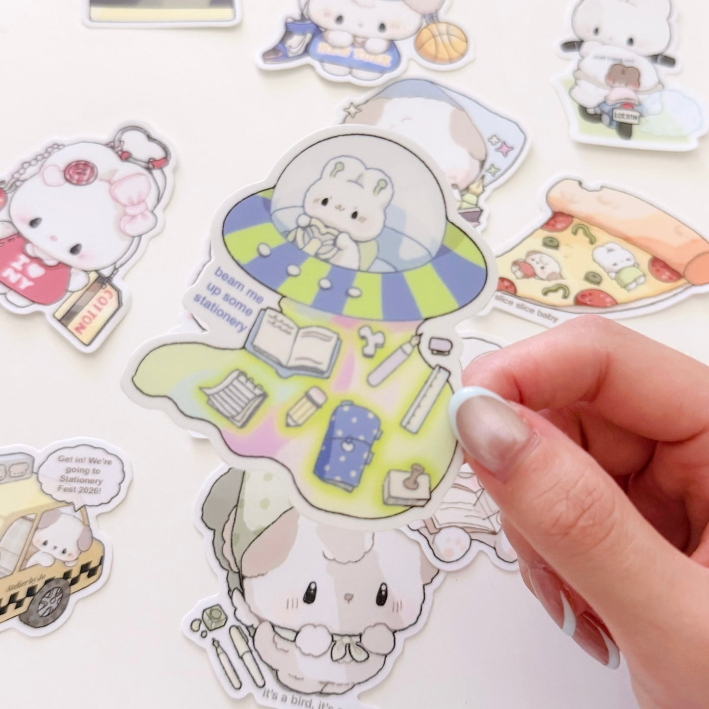 A hand peels a die-cut sticker from a sheet of cute cartoon animal stickers on a white background. The main sticker shows a white rabbit in a green and blue space capsule with books and stationery inside, while other stickers around it show similar cartoon characters in different scenes.