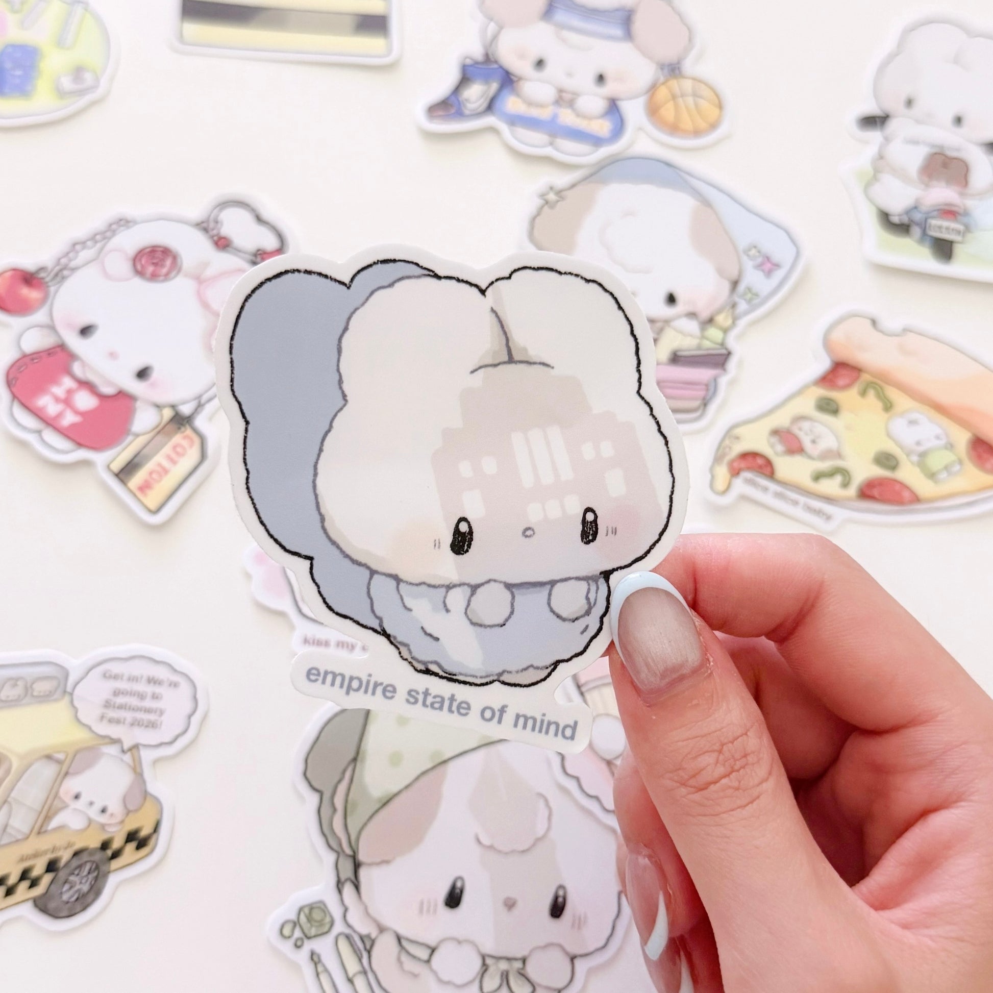 A hand holds a die-cut sticker featuring a cute chibi character with a gray and white cloud-like head, with other colorful stickers scattered in the background on a white surface.