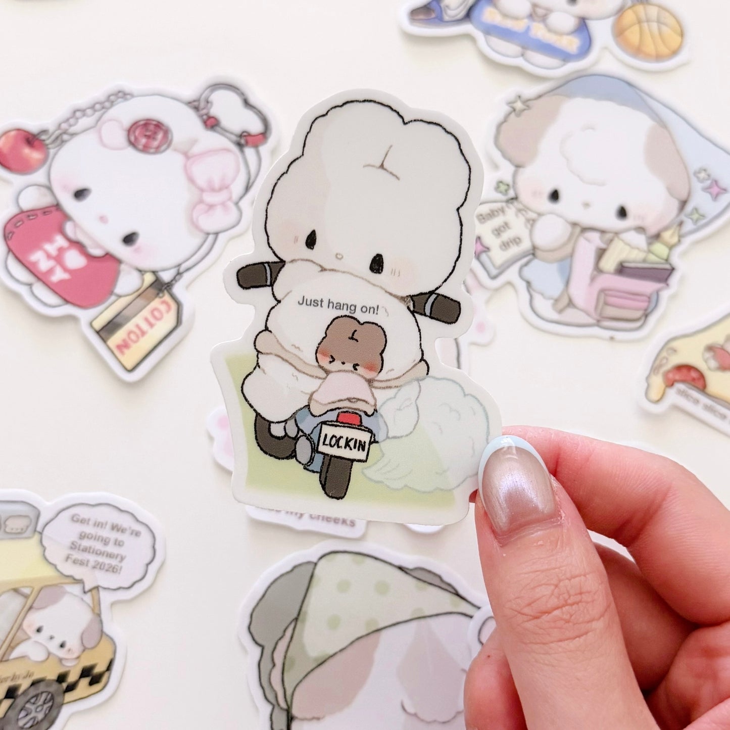 A hand holds a die-cut sticker featuring a cute white sheep character riding a small brown scooter with the text 'Just hang on!' and 'LOCKIN''. Other cartoon stickers are scattered around on a light background.
