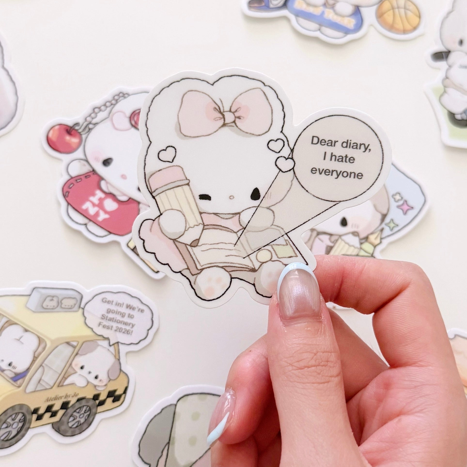 A hand holds a die-cut sticker featuring a cute white bunny character with a pink bow, holding a mug and a speech bubble that reads 'Dear diary, I hate everyone'. Other cartoon stickers are scattered in the background on a light surface.