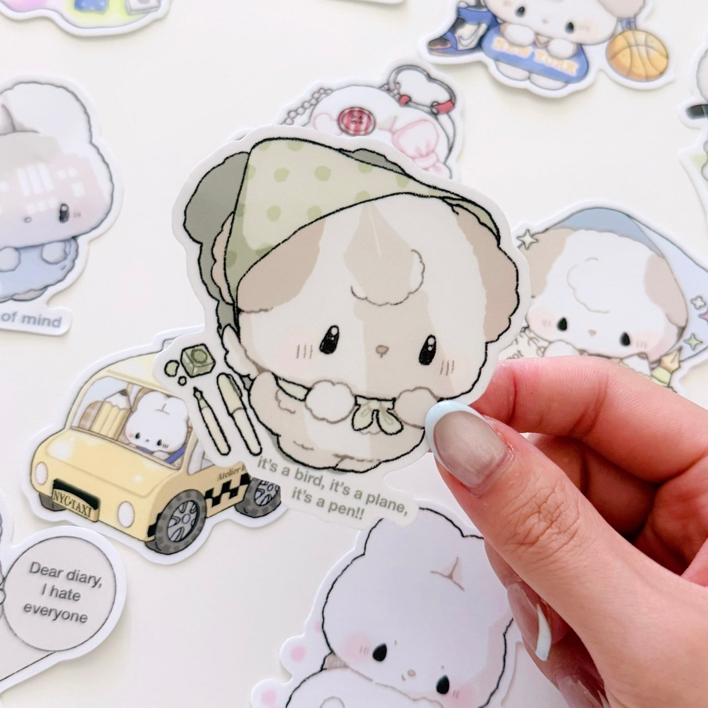 A hand holds a cute die-cut sticker featuring a small white bunny wearing a beige polka-dot bonnet, surrounded by other pastel cartoon stickers on a white background.