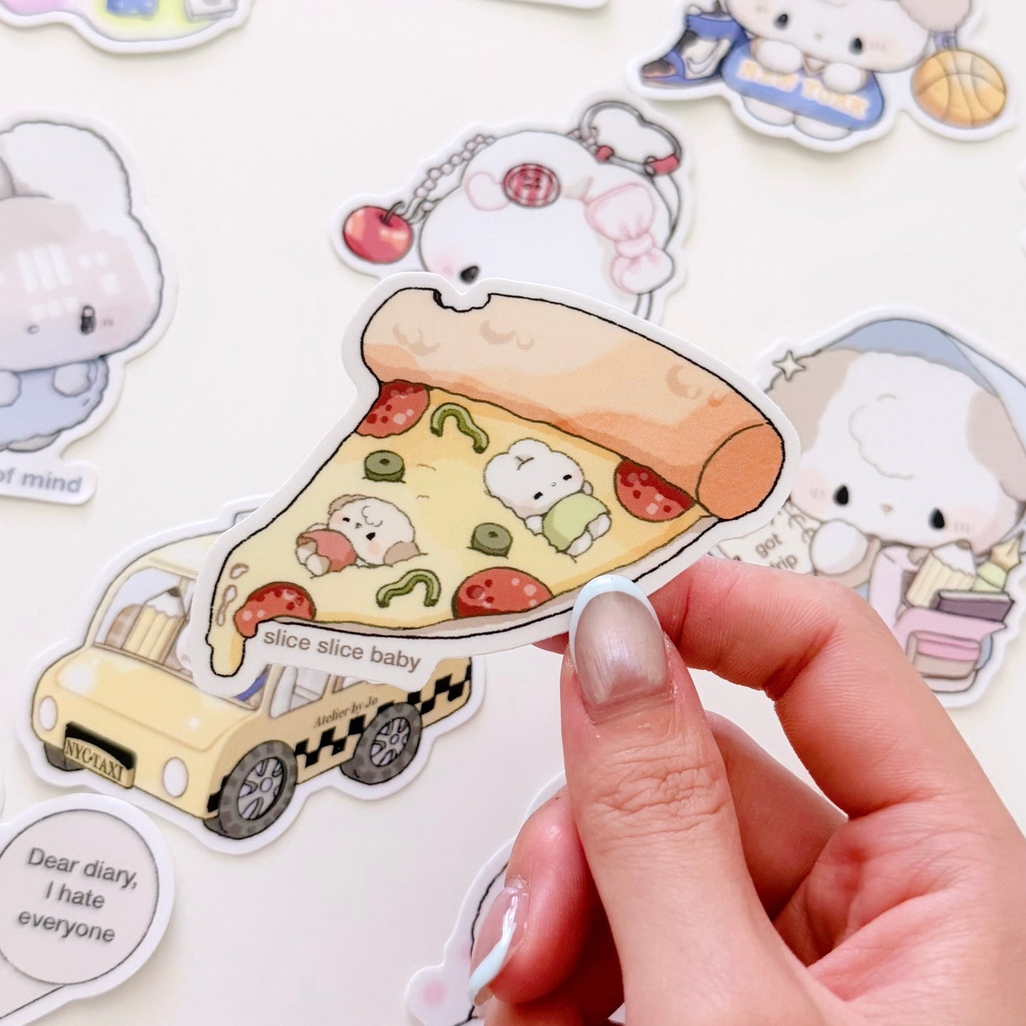 A hand holds a die-cut sticker featuring a slice of pizza with cute cartoon characters, surrounded by other pastel-themed stickers on a white background.