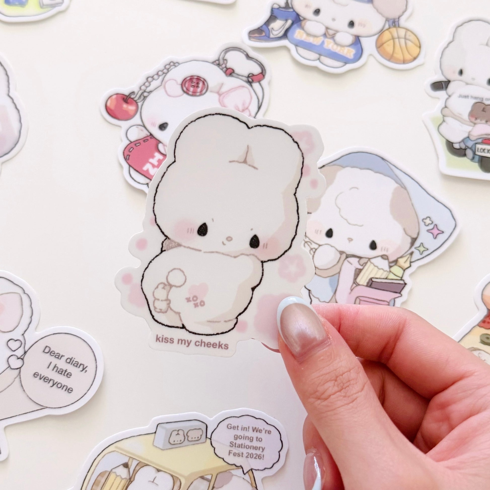 A hand holds a die-cut sticker featuring a cute cream-colored bunny character with the text 'kiss my cheeks' on a white background, surrounded by other pastel cartoon stickers.