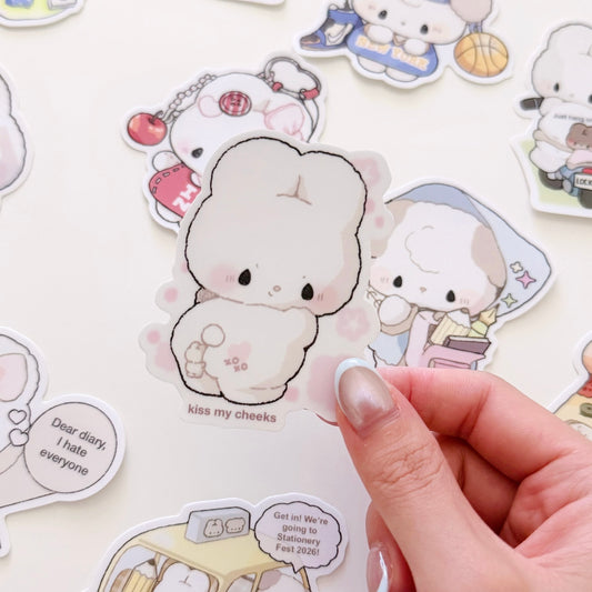 A hand holds a die-cut sticker featuring a cute cream-colored bunny character with the text 'kiss my cheeks' on a white background, surrounded by other pastel cartoon stickers.