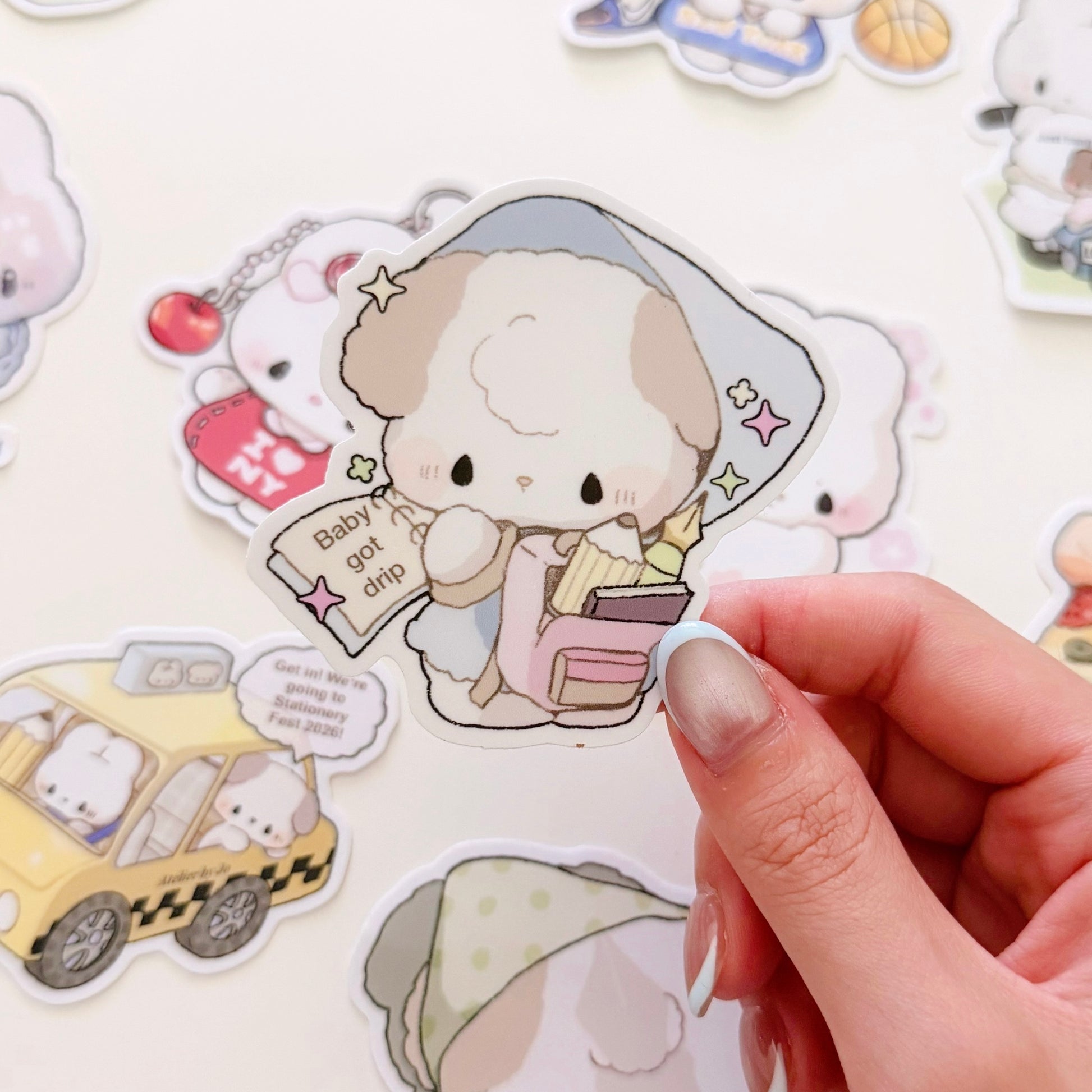 A hand  holds a die-cut sticker featuring a cute cartoon dog character wearing a blue-gray nightcap and holding a book, with the text 'Baby got grip'. Other cartoon stickers are scattered on a white surface in the background.