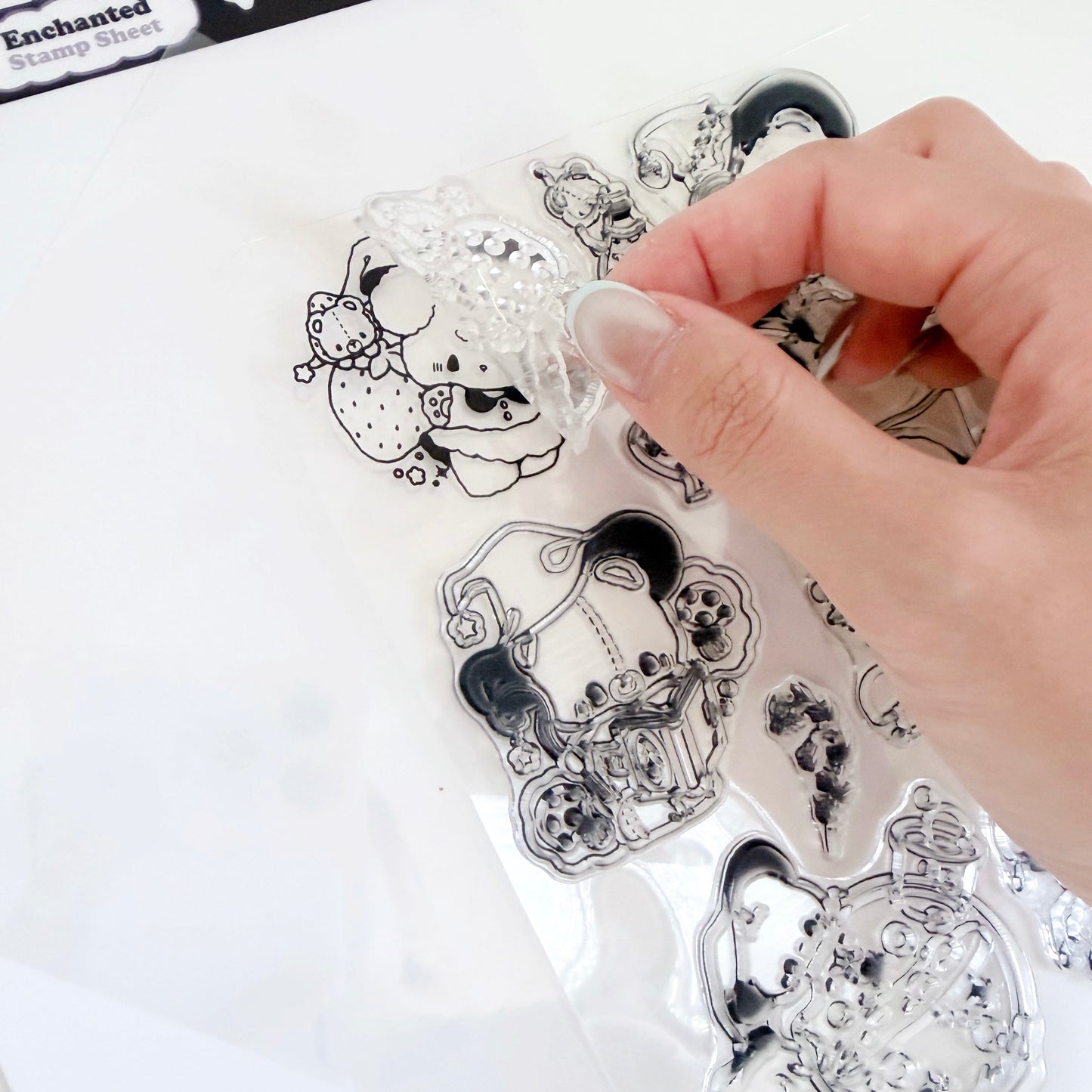 A hand is holding a clear plastic sheet of black-and-white illustrated stamp designs over a white surface. The sheet includes whimsical character and animal outlines, and part of a package label reading 'Enchanted Stamp Sheets' is visible at the top left.