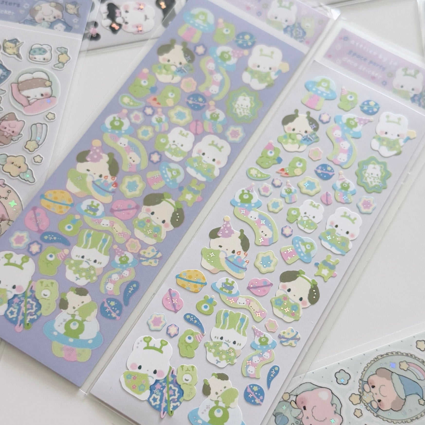 Three packaged sheets of cute cartoon stickers laid out on a white surface. The stickers feature pastel-colored characters and motifs including bunnies, frogs, flowers, stars, and small illustrated objects in soft purple, green, pink, blue, and white tones.