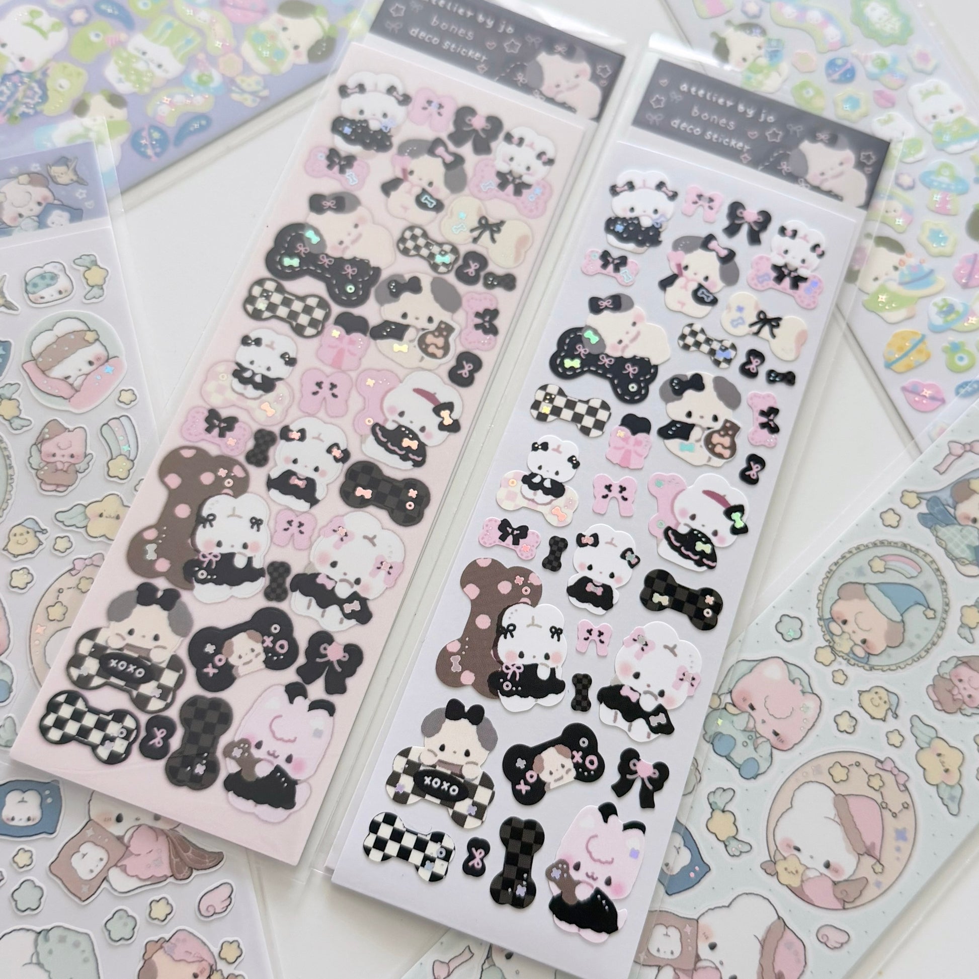 Two packages of cute cartoon character stickers, featuring black-and-white and pink-and-white designs, displayed on a white surface with other sticker sheets partially visible.
