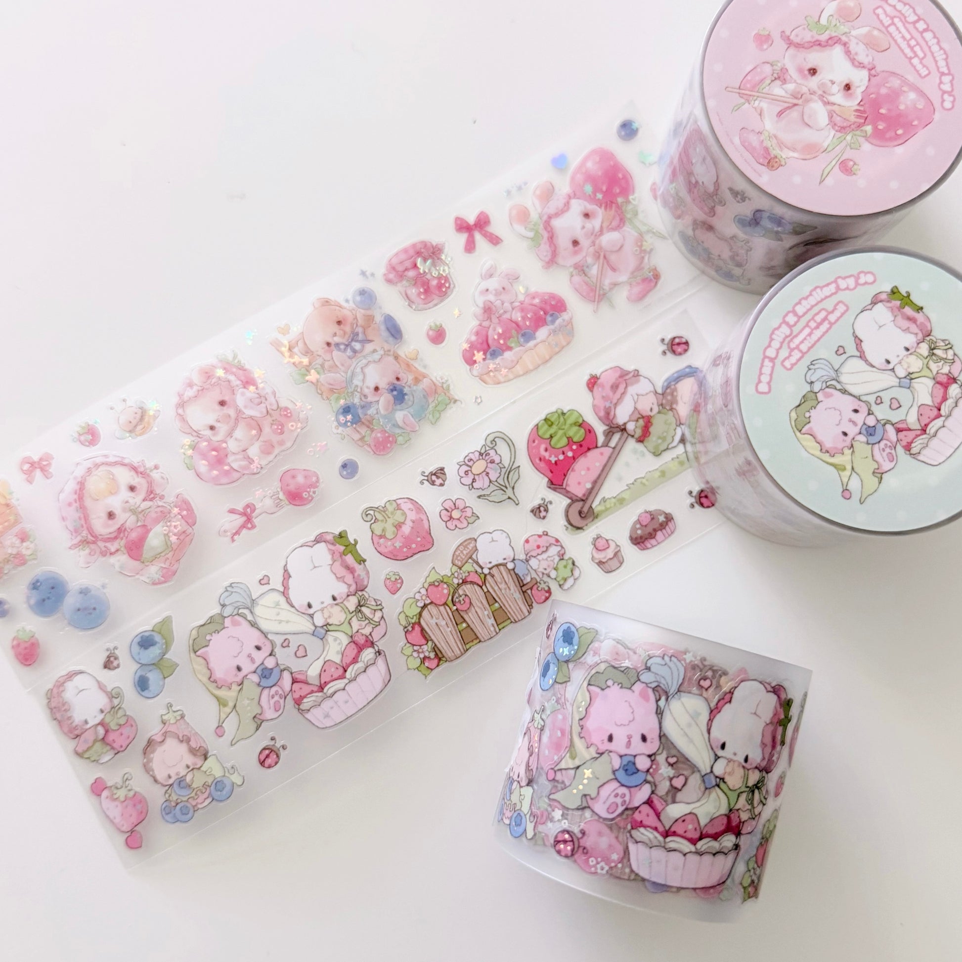 A set of cute pastel washi tape products on a clean white background, including a long unrolled strip and three rolled tubes. The designs feature cartoon characters, strawberries, cupcakes, and small decorative motifs in soft pink, blue, and green tones.