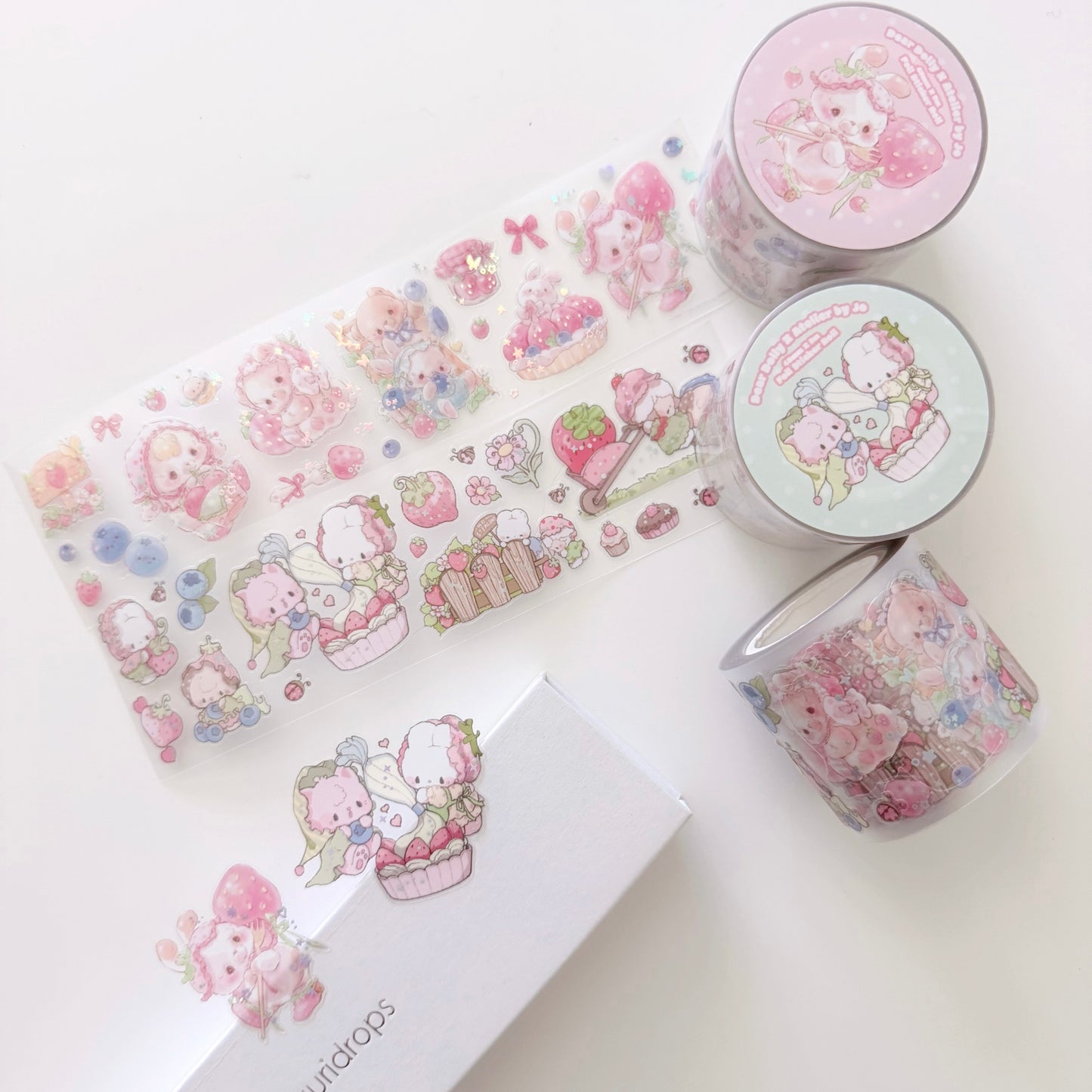 A flat lay of cute pastel washi tape products on a white background, including a long decorative tape strip, two round rolls, one unrolled section showing the pattern, a branded white box, and a small card. The designs feature pink cartoon characters and floral/cupcake motifs.